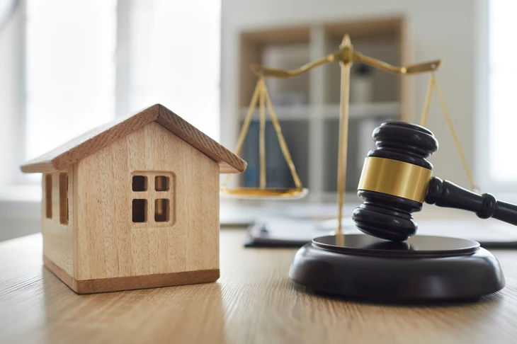 Scène d'intérieur lumineux avec bureau en bois présentant une maquette de maison en bois naturel, un marteau de juge en ébène avec bande dorée sur socle circulaire, et une balance de justice dorée en arrière-plan