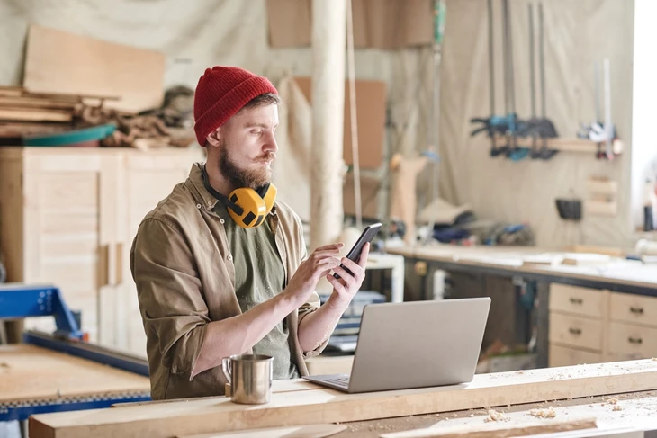 Artisan barbu en bonnet rouge travaille dans atelier menuiserie, consulte smartphone près ordinateur portable ouvert sur établi bois