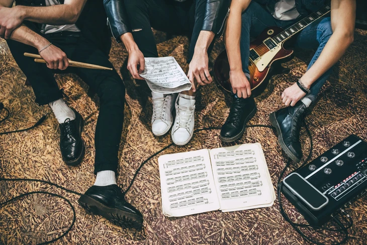 Quatre musiciens assis en cercle sur un tapis à motifs, travaillant sur des partitions musicales lors d'une session de répétition ou composition collaborative en intérieur