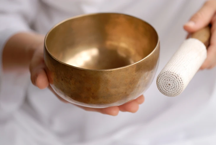 Personne en vêtement blanc tenant un bol chantant tibétain doré et un maillet dans un environnement intérieur calme et lumineux